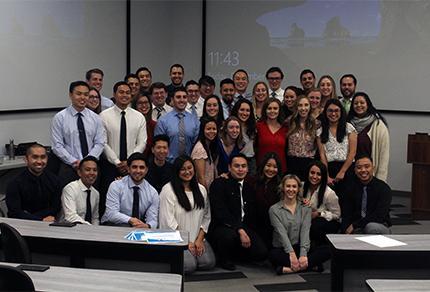 A group of students in business attire take a group photo at the front of a lecture hall