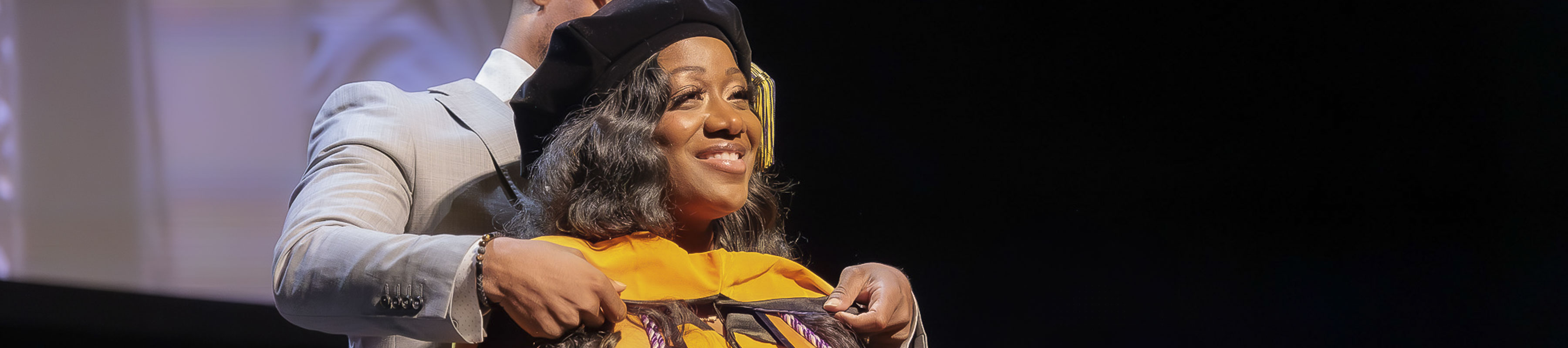 A doctoral hood is placed on a graduating student on stage during the commencement ceremony.