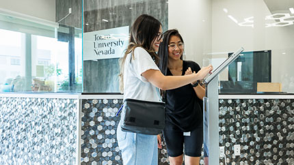Two students enter information into a tablet on a stand within an glass enclosed lobby.