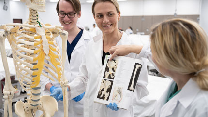 Two students and a faculty member, all wearing white coats, compare images in a textbook to a medical skeleton in a classroom