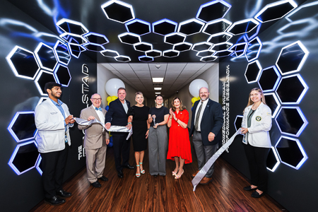 Representatives from Engelstad and Touro University Nevada stand in a long hallway under a lighted sign that says The Engelstad Research Complex. The woman in the center of the group cuts a satin ribbon with an oversized pair of scissors.