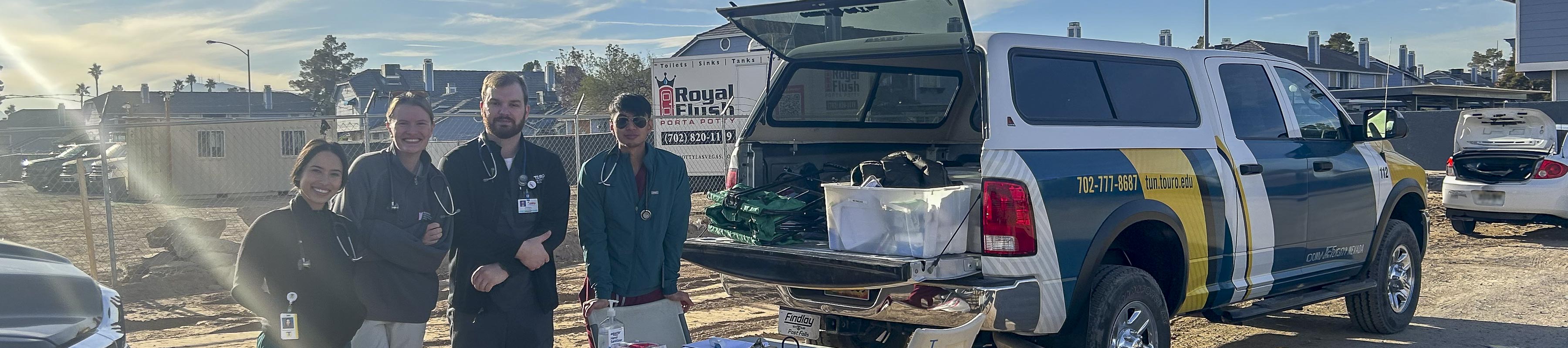 Four students wearing stethoscopes stand near a folding table and a truck bed filled with medical supplies in a dirt parking lot