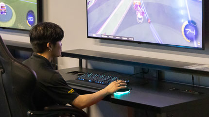 A young student sitting at a computer desk playing a car racing game on large monitor.