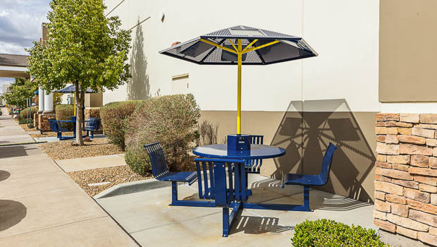 Two outdoor metal solar tables with umbrellas and four attached chairs sit in paved coves along the side of the building\'s exterior.