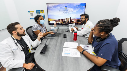 Four students talking at a table in a private study room with a TV monitor behind them.