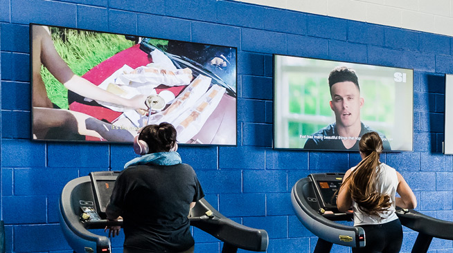 Two students walk on treadmills while watching different programs on two large flatscreen televisions that are hung on the wall in front of them.