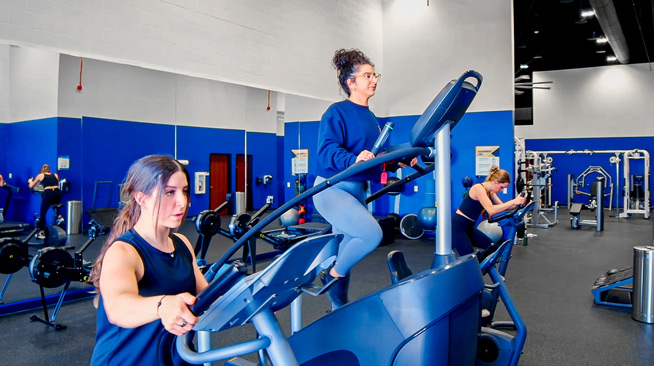 Three students workout on cardio machines. One student is using stationary bike, one a star climbing machine, and the third is on an elliptical.