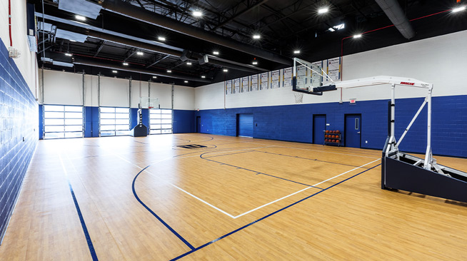 An indoor court with windows containing two basketball hoops on either end, and a rack of basketballs along the back wall.